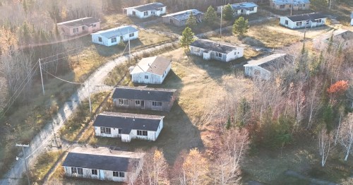 Abandoned buildings in a grassy area under a clear blue sky, with power lines and scattered trees in the background.