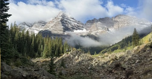 Snowy mountains in the background with clouds partially obscuring the mountains. Conifer trees in the midground; rocks in the foreground. A beautiful mountain vista.