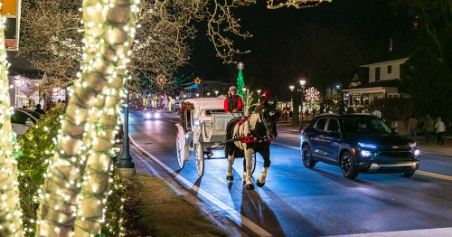 A horse drawn carriage drives on a street at night with Christmas lights next to a car.