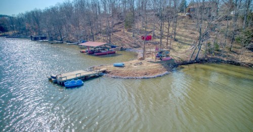 A tan wooden pier/deck on a greenish lake beside a small blue watercraft at the bottom of a hill