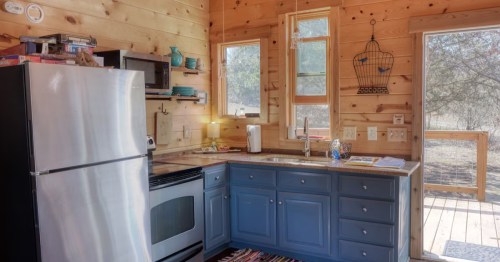 A cabin kitchen with shiny silver fridge, blue cabinets and bare tan wooden walls