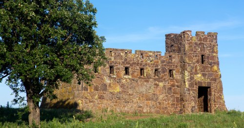 The reddish sandstone walls of Coronado Heights against a pale blue sky and beside a green tree
