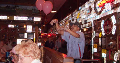 A man in a striped blue and white button down shirt with red suspenders and blue hat throws a roll across the dining room at Lambert's