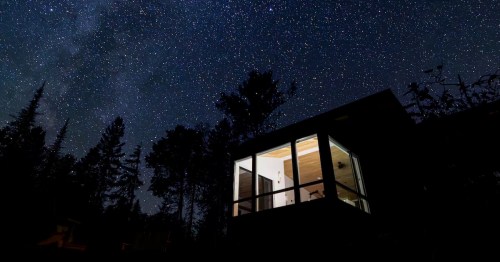 Lights from a modern cabin in a forest are visible against a dark, starry sky.