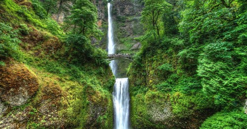 A tall, thin waterfall drops dramatically down a green cliffside. A bridge sits across it.