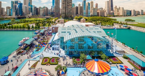 A view of Navy Pier from above with bright green and red plants, an orange carousel and blue pier building covered in glass with the city of Chicago in the background