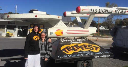 Two teens stand in front of a replica of the Star Ship Enterprise on a float labeled "TrekFest" beneath a blue sky.