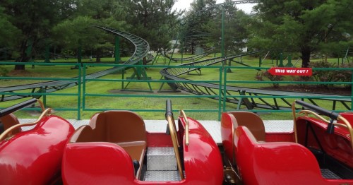 the red cars of a rollercoaster in the foreground and the tracks hovering above green grass at santa's village in new hampshire