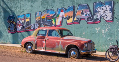 a classic yet beat-up red car sits in front of a colorful mural with the word seligman painted on a wall