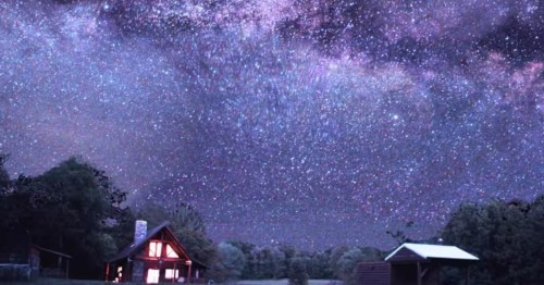 Extreme night sky shot showing the Milky Way over a small, lighted cabin and outbuildings. 