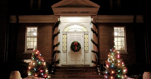 the brick exterior of the stockbridge library with green garland decorating its white columns and two trees with colorful christmas lights in stockbridge, massachusetts