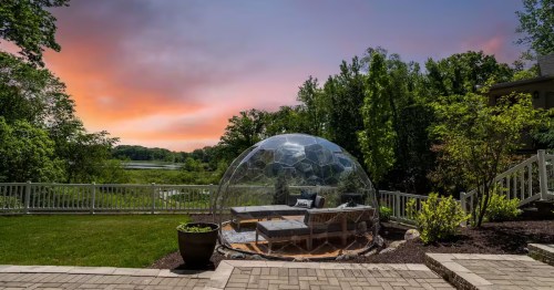 A clear outdoor dome with seating is set against a vibrant sunset sky and a backdrop of green trees and a lake.