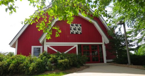 A red barn-style building surrounded by trees and parked cars under a clear blue sky.