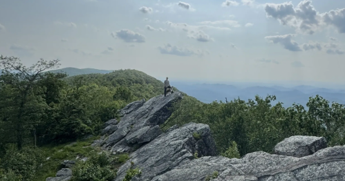 Hiker stands on rocky overlook, with green and blue mountains in the distance under a partly cloudy summer sky