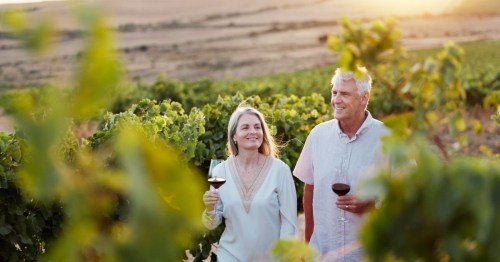 A happy couple strolling through a vineyard while sipping red wine.