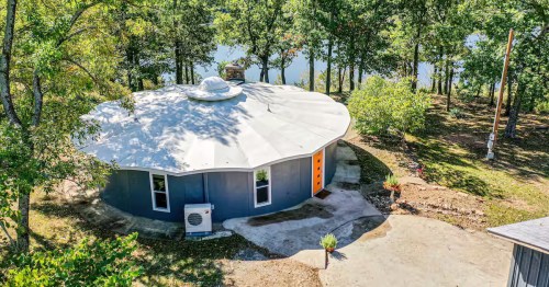 An aerial view of a gray, circular yurt-style house with a white roof nestled among trees near a lake. 