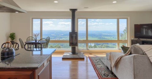 Bright living room with hardwood floors, a central fireplace, and large windows overlooking a stunning valley view.