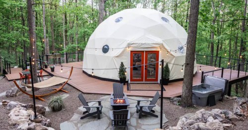 Geodesic dome rental nestled in a forest clearing with a large wraparound deck, hot tub, and fire pit area.
