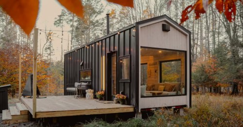 Exterior view of a modern black and light wood tiny home with string lights and a deck in an autumn forest.