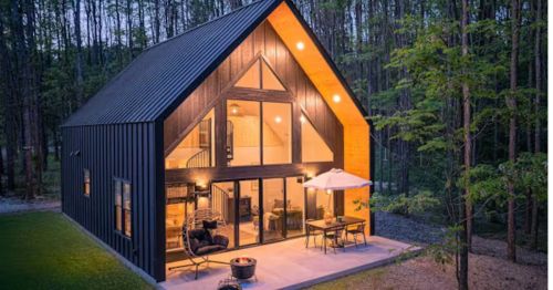 The exterior of a well-lit modern cabin in the woods with large glass windows.