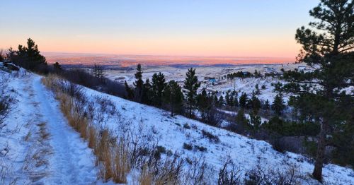 A snowy hillside trail overlooks a rolling valley of scattered evergreens and distant city lights under a soft pink and blue sunset.