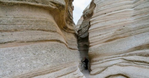 Traveler standing between steep, curving slot canyon walls with distinct volcanic tuff layers at Kasha-Katuwe Tent Rocks National Monument, New Mexico.
