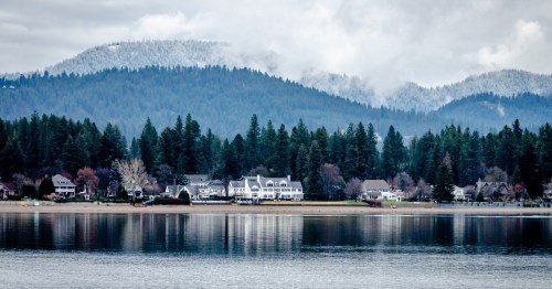 Luxury homes border a lake with tree-laden hills shrouded in clouds in the background.