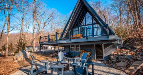 Charming vintage black A-frame cabin nestled in a wooded setting, featuring a deck and a fire pit area below.