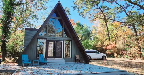 Stylish dark A-frame cabin with large glass front windows, a grey deck, and Adirondack chairs set in a wooded area.