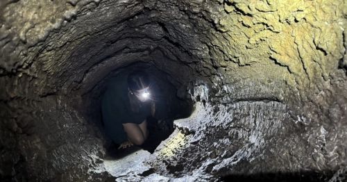 A person wearing a headlamp crawls through a narrow, textured cave tunnel, partially illuminated by their light.