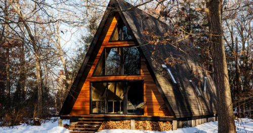 Wood-paneled A-frame cabin with large glass windows and a dark roof, surrounded by winter woods and snow.