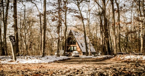 A-frame cabin partially visible down a dirt road, set deep in the winter woods with scattered snow.