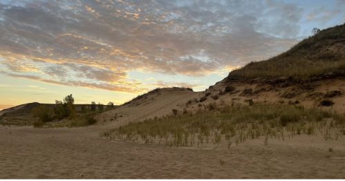 A sand dune and sandy beach with the sun setting behind it.