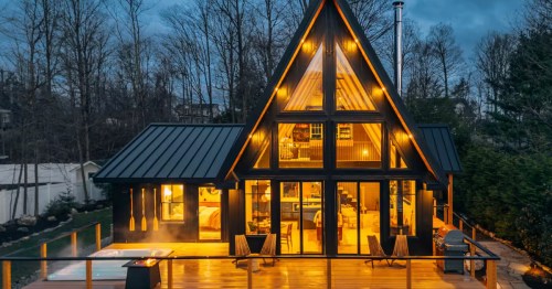 Striking black modern A-frame cabin with massive glass windows, a hot tub, deck, and fire pit at twilight.