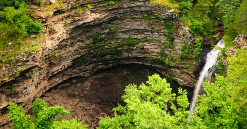 Overlook view of Cedar Falls at Petit Jean State Park in Arkansas, showing a tall waterfall pouring into a massive rock amphitheater surrounded by lush green forest and layered sandstone cliffs.