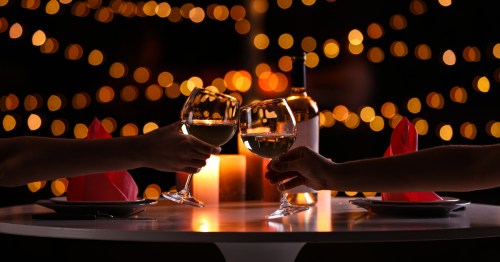Two people clink wine glasses across a table with twinkling lights in the background.