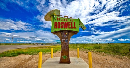 Colorful roadside ‘Welcome to Roswell’ sign with a green UFO-themed design and red lettering standing in an open desert landscape.