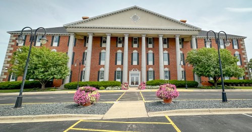 A brick mansion with six white pillars and a large parking lots with planters of pink flowers