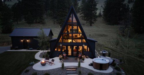 An A-frame Airbnb lit up at night with a hot tub, fire pit, and outdoor chairs outside the rental.
