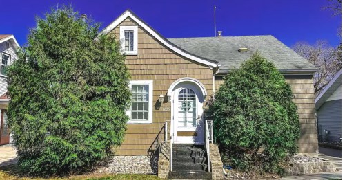 A cute cottage with shingle siding, a white, arched front door, and a peaked roof surrounded by big, green bushes and trees.