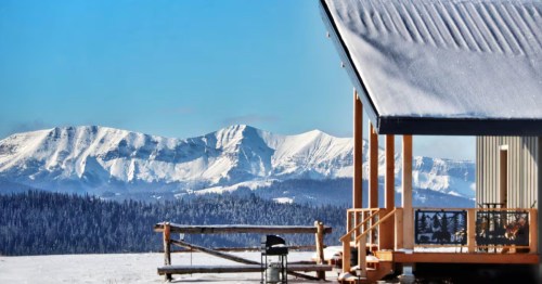 The side of a cabin vacation rental is visible in front of a bold mountain range and a pine tree forest covered in snow.