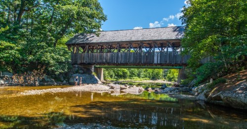 A covered bridge rests over a shallow brown river with greenery on either side.