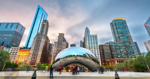 A pink sky on the left and cloudy, dark sky on the right behind a row of downtown Chicago skyscrapers. The silvery Cloudgate sculpture is in the center foreground.