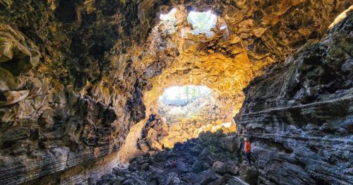 A hiker standing inside a massive lava cave with two large openings above at a hidden gem park in New Mexico.