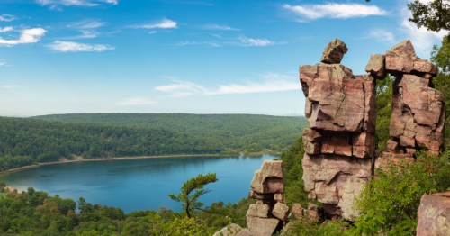 A bright blue lake surrounded by green trees in the background under a blue sky. A large, tan rock formation that looks like a doorway on the right side.