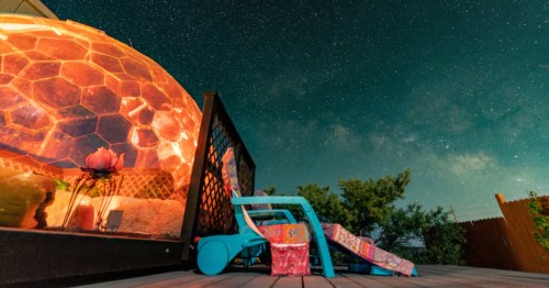 two louonge chairs sit outside a dome home aglow at night under the Milky Way