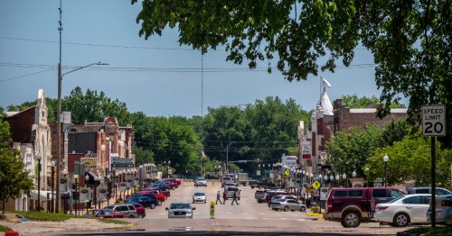 View down a wide street in historic Council Grove, KS, showing parked cars and two people crossing the road.