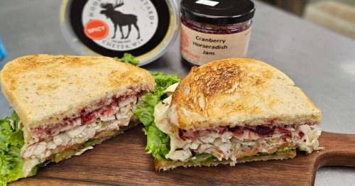 A turkey sandwich with lettuce and other ingredients on toasted sourdough rests on a dark wood cutting board near jars of mustard and cranberry sauce.