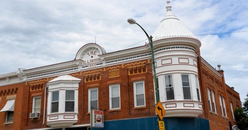A white turret and dormer windows grace a historic red brick building under clouded blue skies.