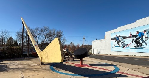 The world's biggest hockey stick and puck stand next to a colorful mural of boys playing hockey on a frozen pond.